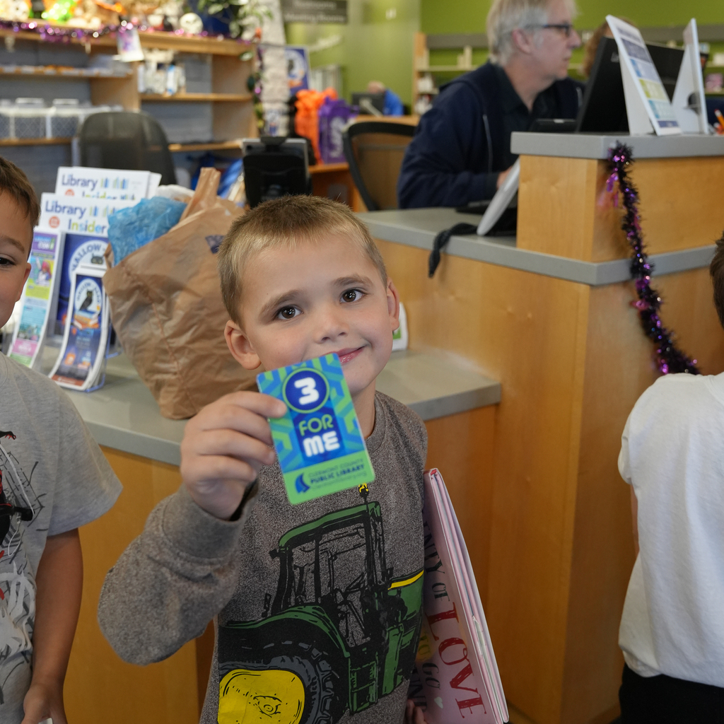 A student smiles and displays a “3 for Me” library card while holding books at the checkout counter.