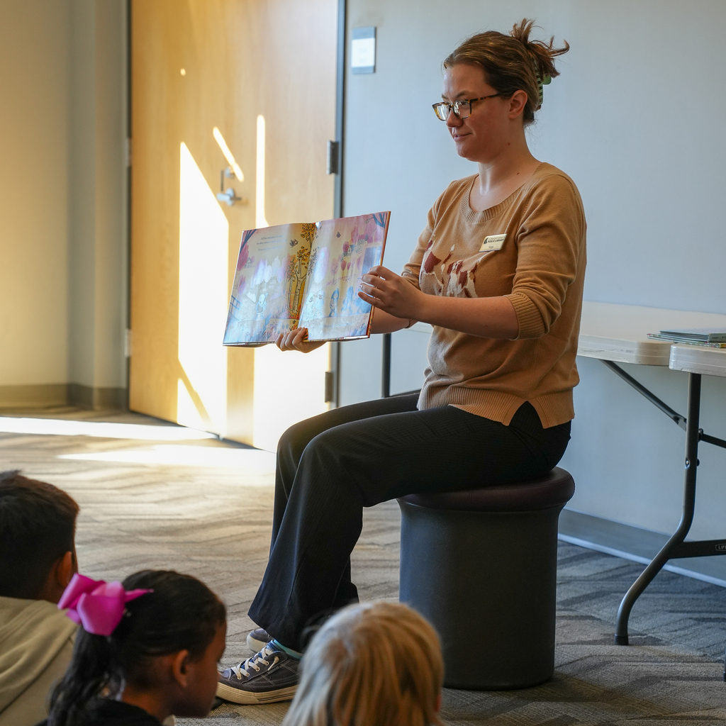 A library staff member holds an open picture book toward kindergarten students seated on the floor.