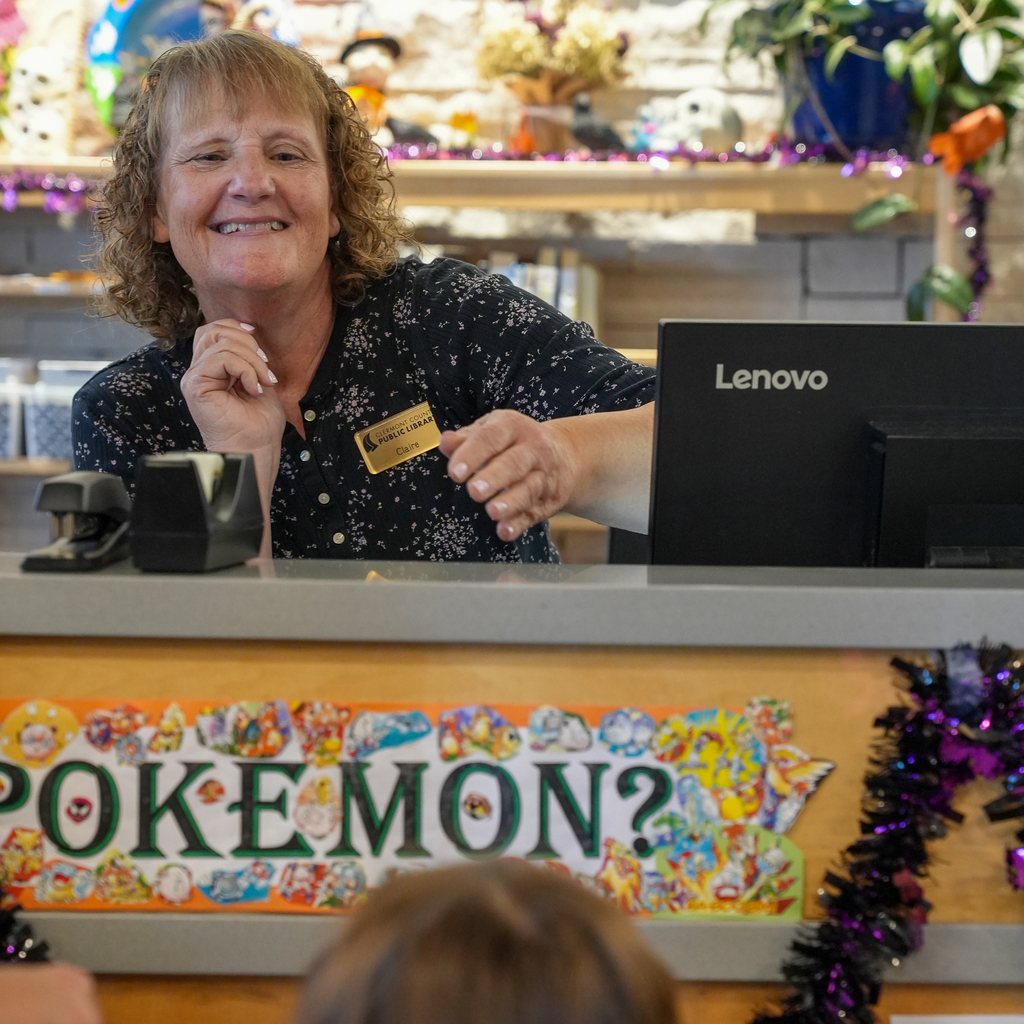 A library staff member smiles behind the checkout counter, decorated with Pokémon characters and Halloween garland.