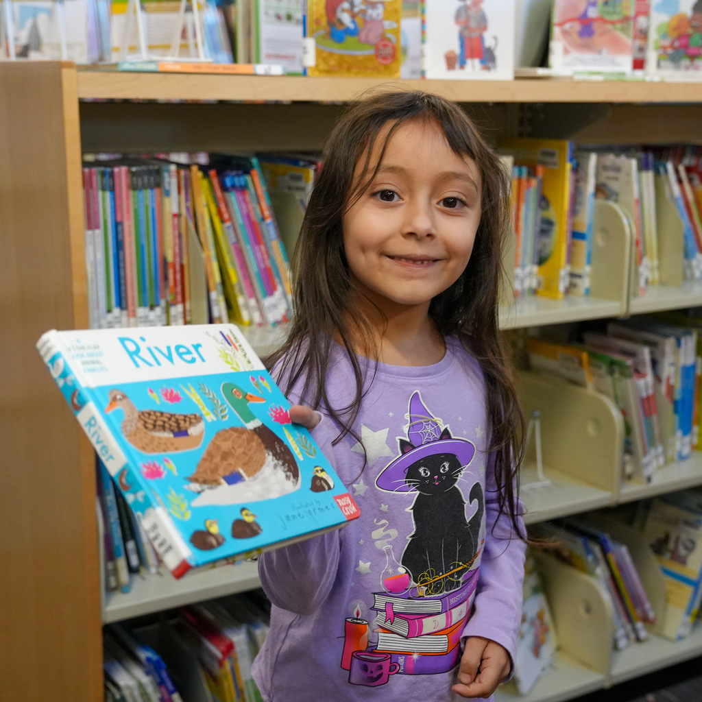 A student holds up a book titled “River” while standing in front of shelves of children’s books.
