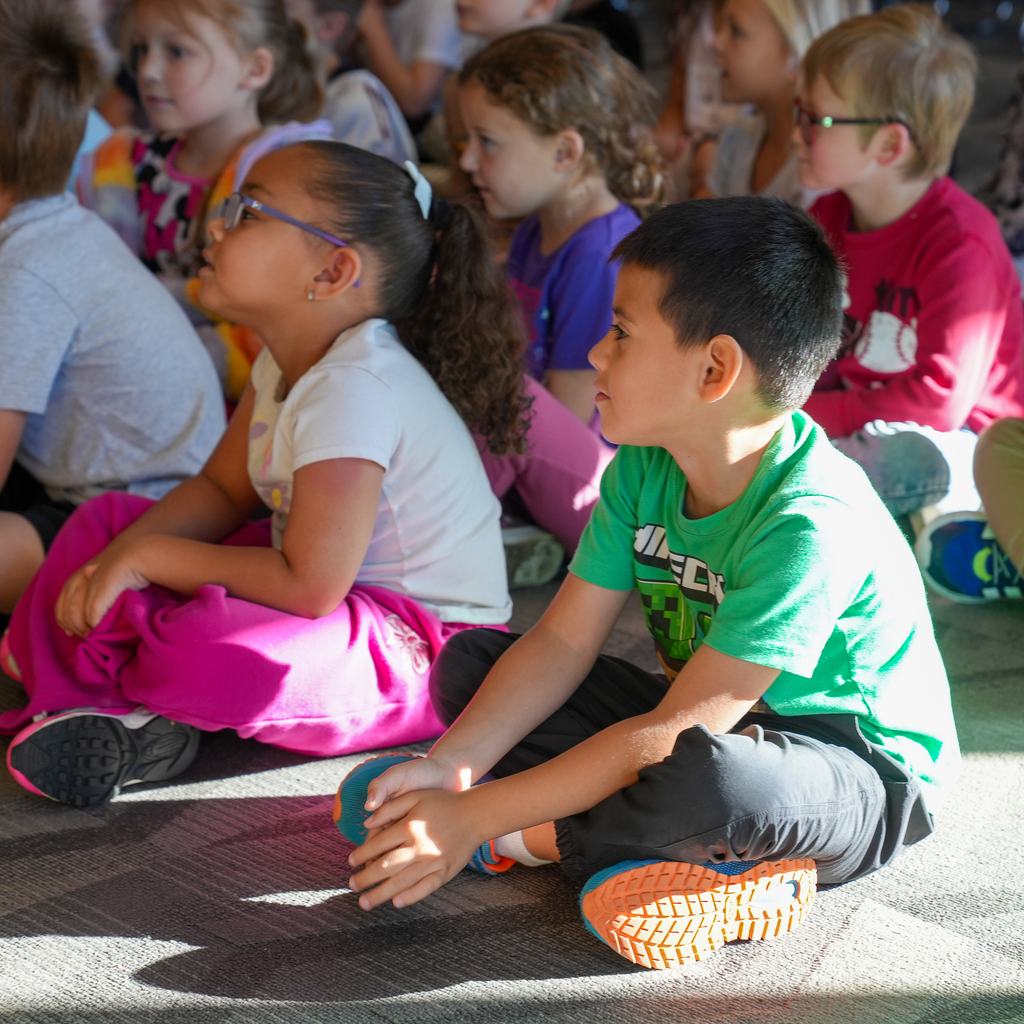 Mulberry kindergarten students sit cross-legged on the library floor, looking toward a presenter outside the frame.