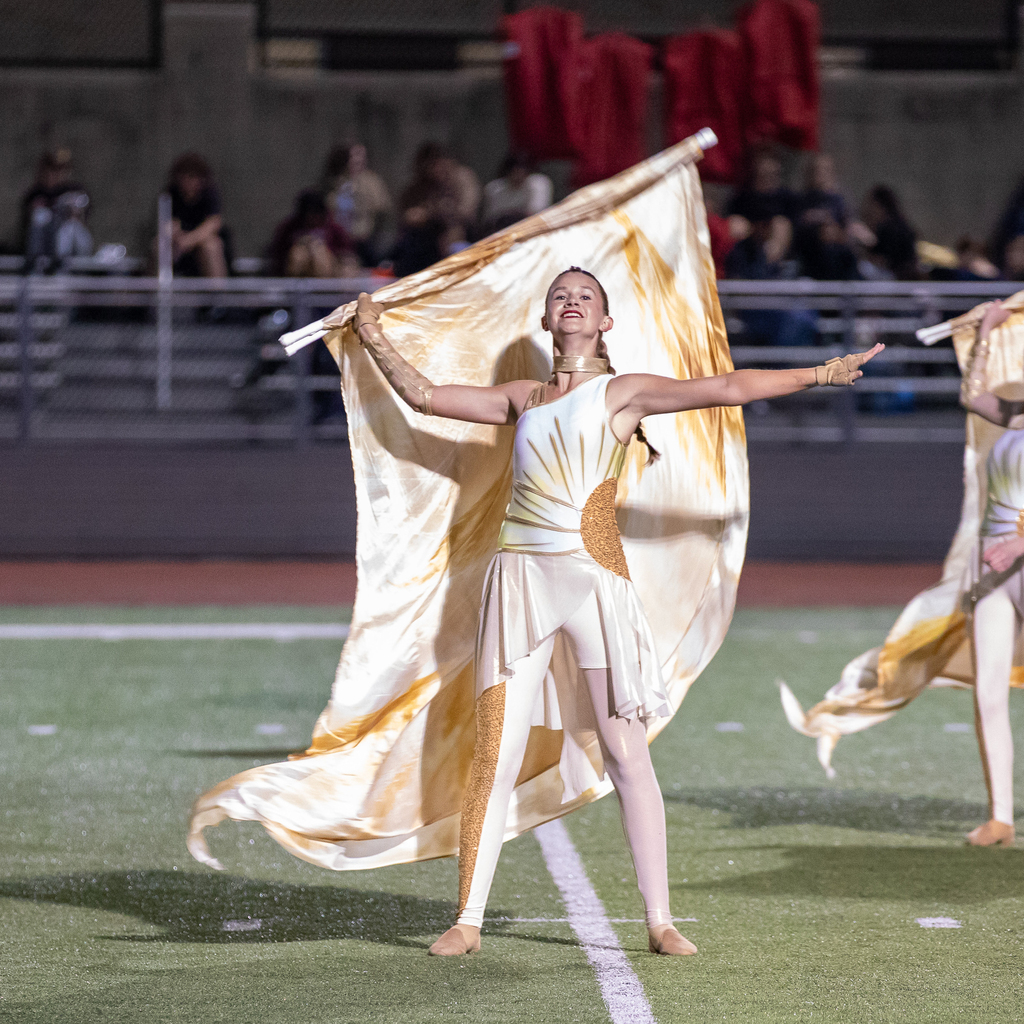 A smiling color guard member extends one arm outward while holding a flowing gold and white flag behind their shoulders.