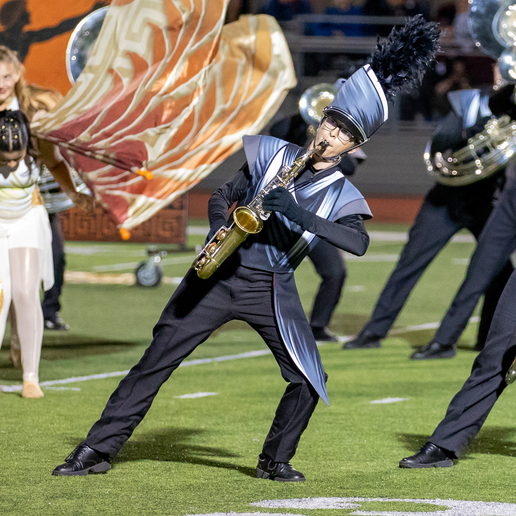 A marching band member plays a gold saxophone while leaning sideways in a wide stance, with flags waving behind them.