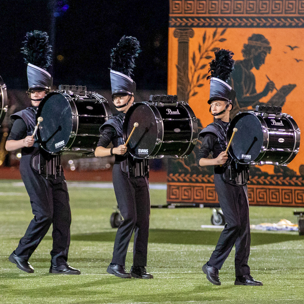 Three bass drummers march across the field in black and silver uniforms with tall feathered hats.