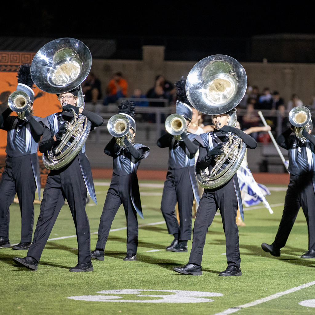Two sousaphone players lead a group of brass musicians performing in formation on the football field.
