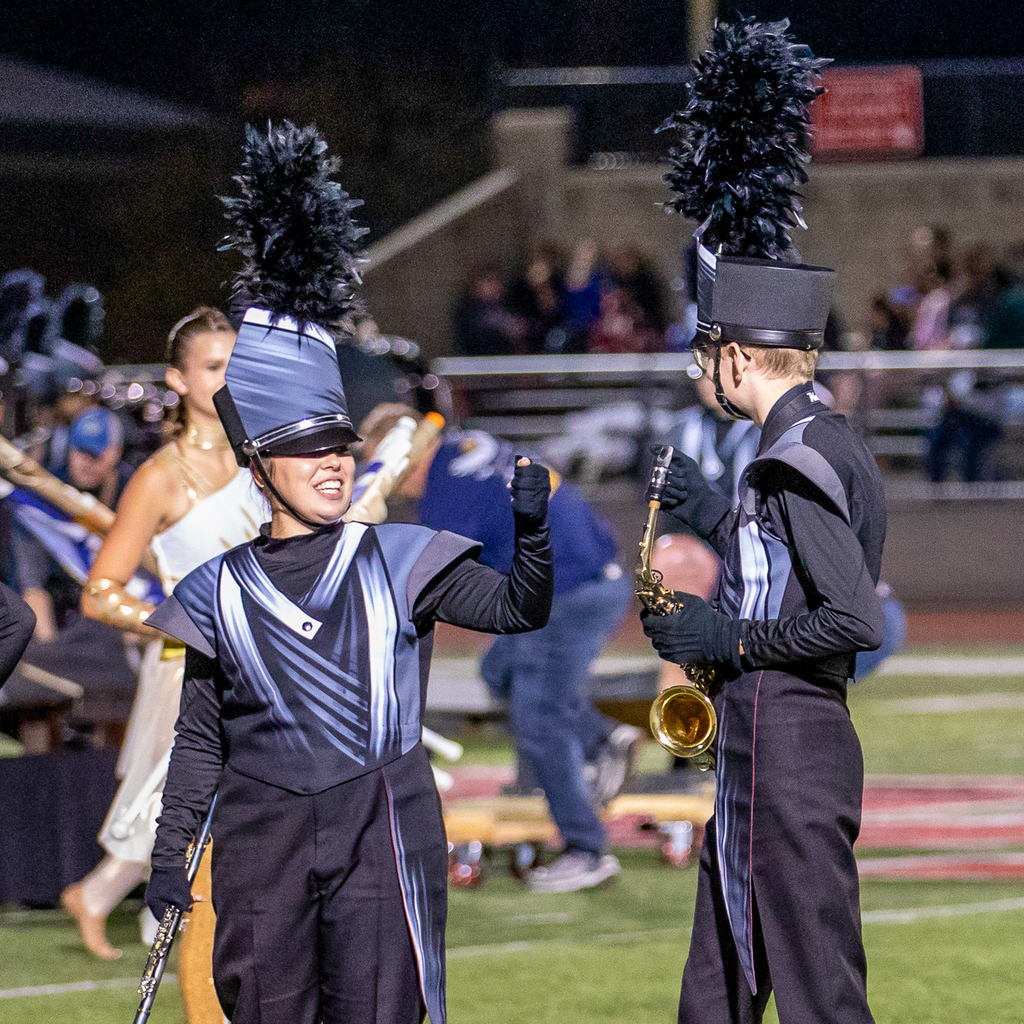 Two marching band members raise their fists toward each other, one smiling with a woodwind instrument and the other holding a saxophone.
