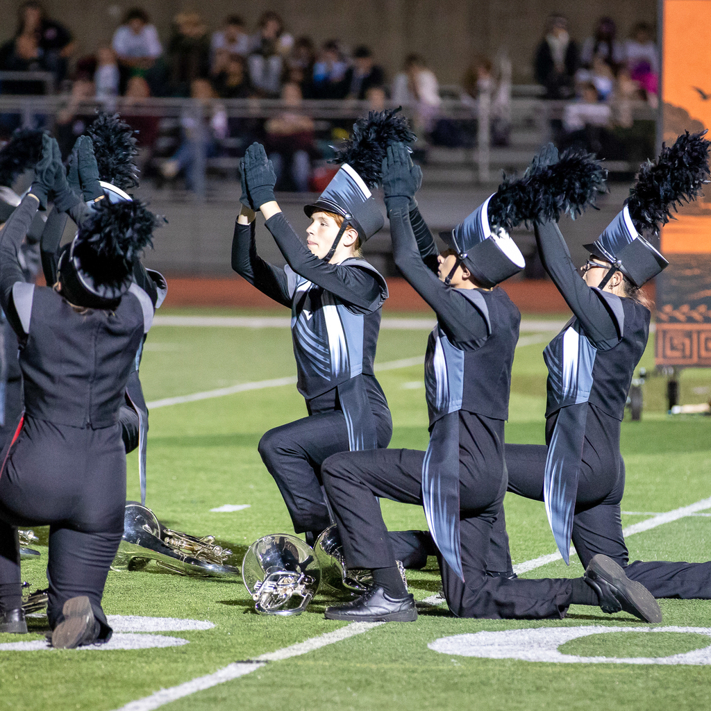A color guard member extends one arm while holding a flagpole across their body, with two teammates performing behind them.