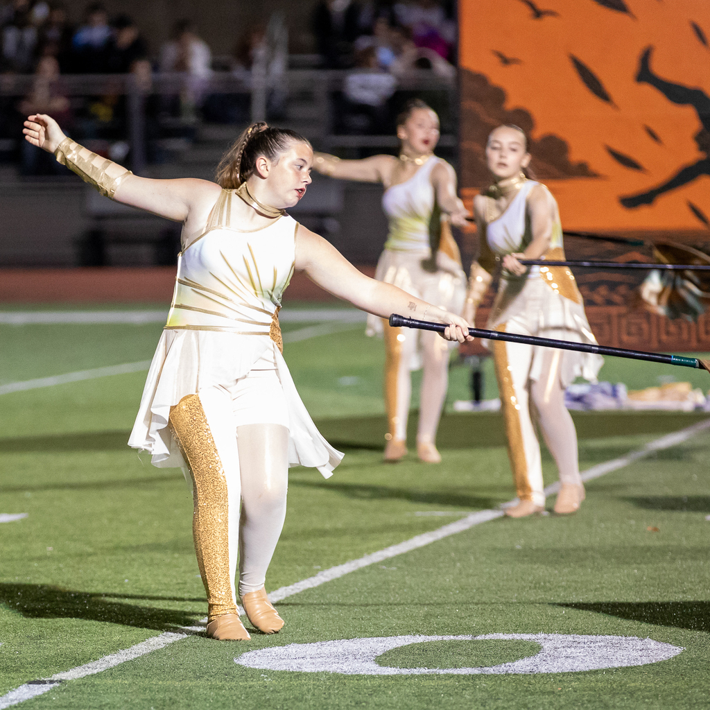 Three color guard members in white and gold costumes sweep patterned flags near the turf, with an orange Icarus backdrop behind them.