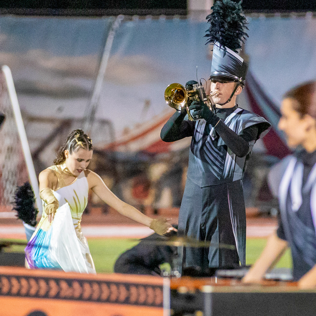 Marching band members kneel on one knee with their gloved hands pressed together overhead and brass instruments resting on the turf.