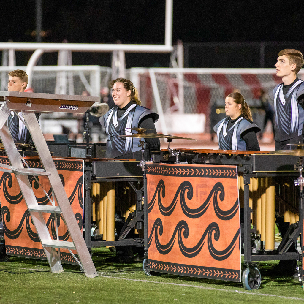 Four percussionists stand behind keyboard percussion instruments and cymbals decorated with orange panels featuring black wave patterns.