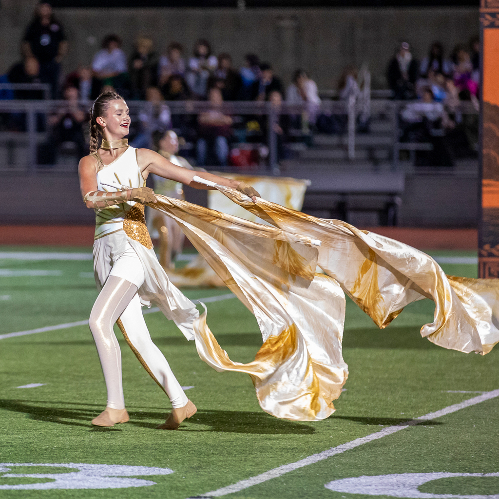 A smiling color guard member in a white and gold costume sweeps two gold and white flags across the football field.