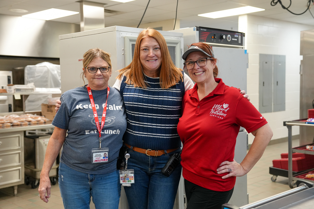 Three smiling staff members stand with their arms around one another in a school kitchen. One wears a red Milford Nutrition Services polo.