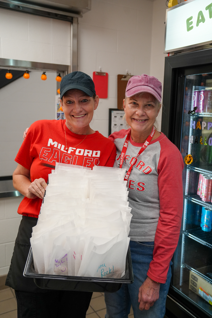Two smiling nutrition staff members in Milford Eagles shirts pose with a tray of individually bagged cookies featuring handwritten messages.