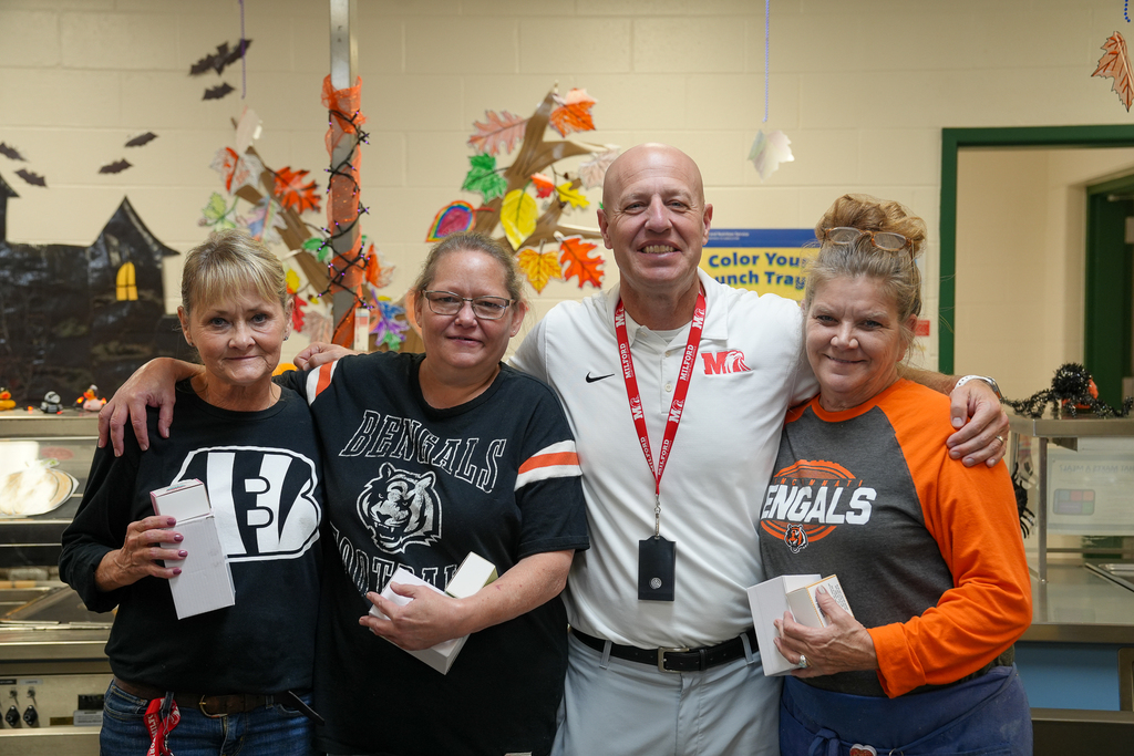 Four smiling staff members pose together in a cafeteria decorated for fall. Three wear Cincinnati Bengals shirts and hold small gift boxes.
