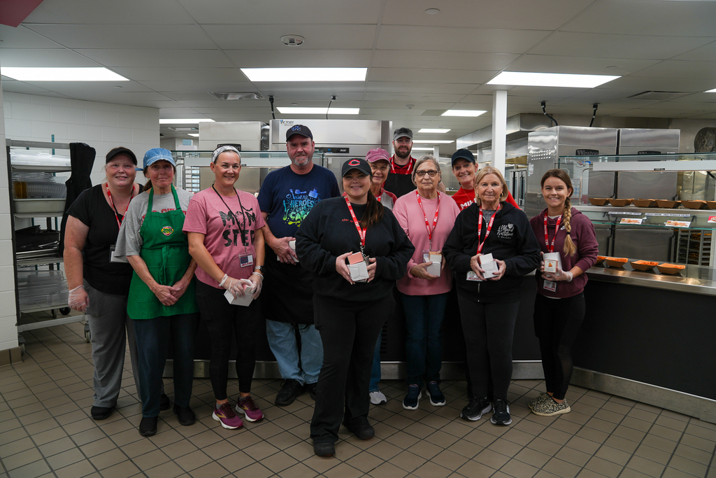 Ten staff members pose together in a school cafeteria kitchen, several holding small gift boxes beside the serving line.