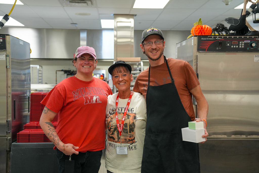 Three smiling nutrition staff members pose together in a school kitchen, with the staff member on the right holding two stacked gift boxes.