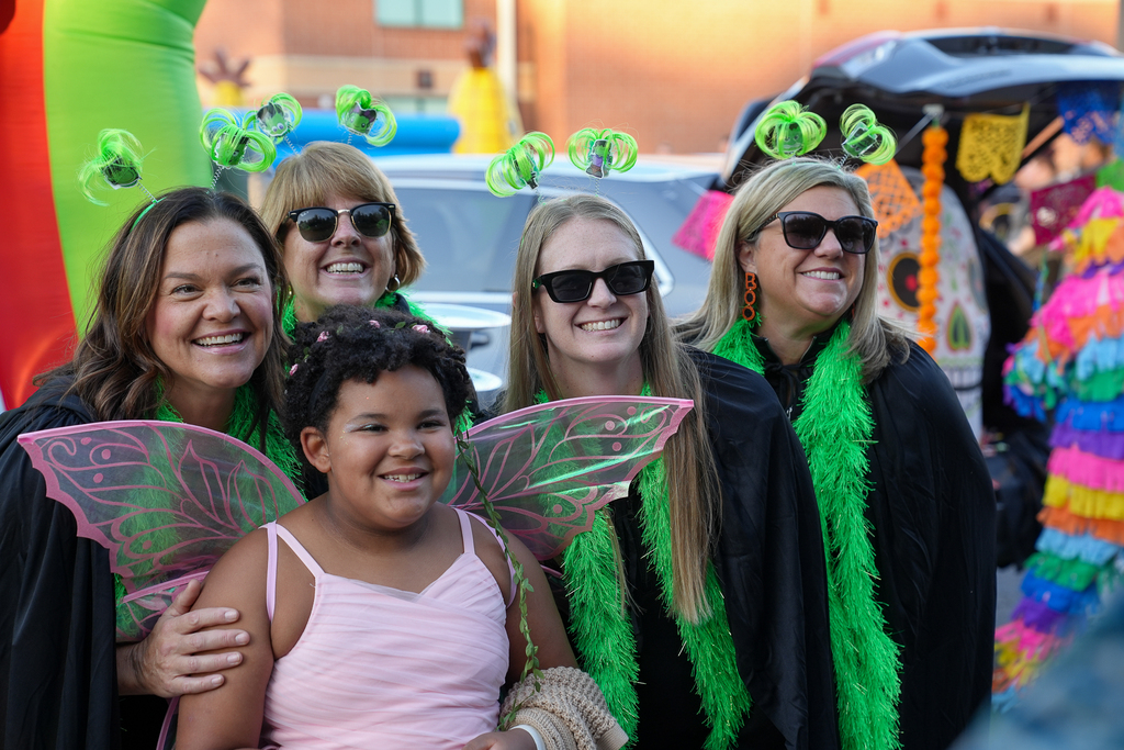 A child wearing pink fairy wings poses with four smiling adults in matching black capes, green feather boas and green novelty headbands.