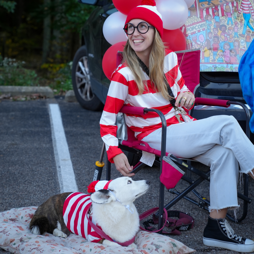 A smiling adult in a Where’s Waldo costume sits beside a dog wearing a matching red and white striped outfit and hat.