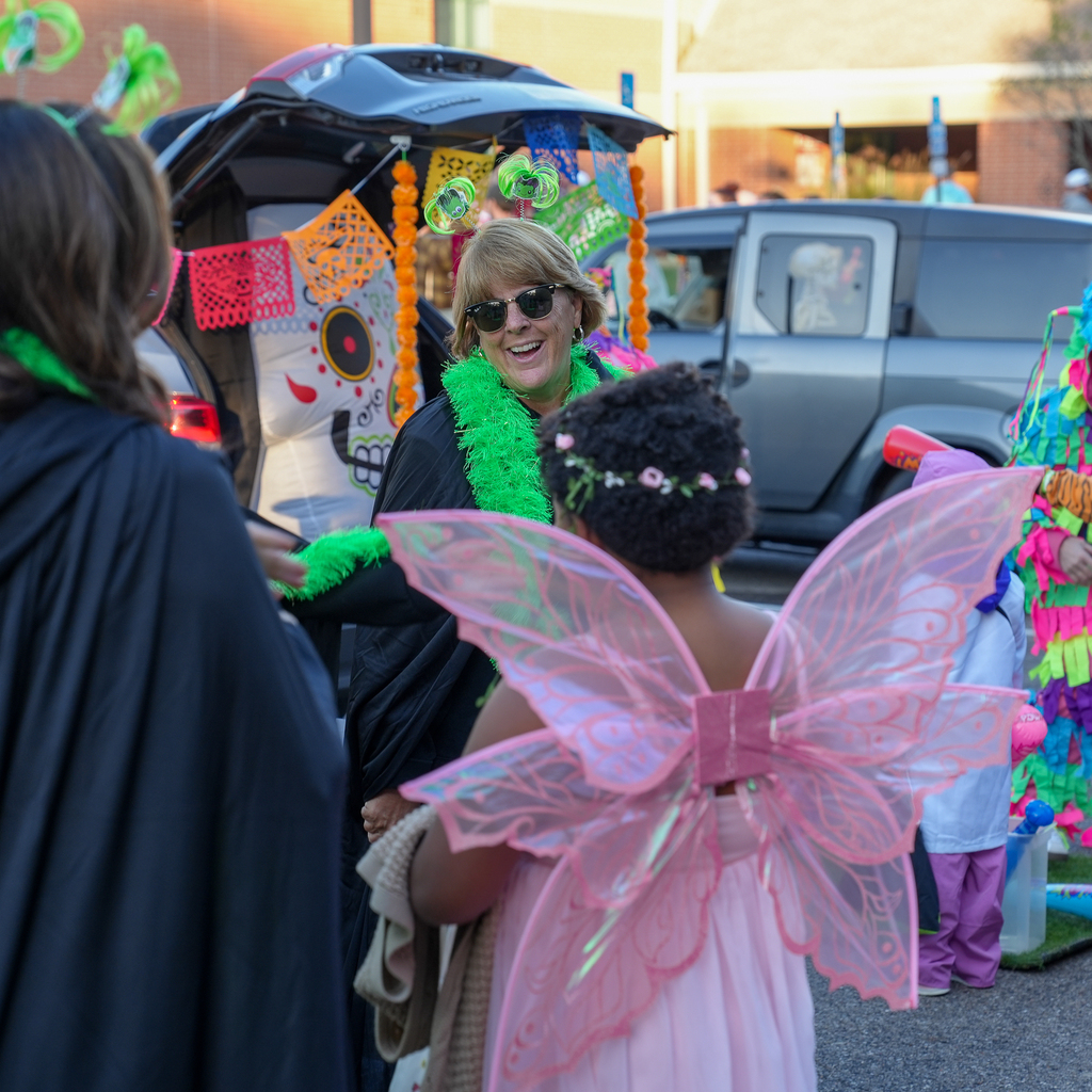 An adult wearing a green feather boa and novelty headband smiles at a child in pink fairy wings beside a decorated vehicle.