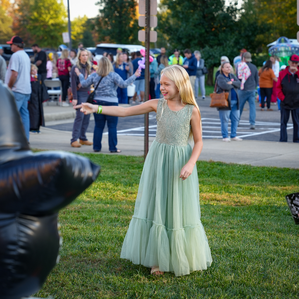 A smiling child in a light green dress extends an arm toward an inflatable game target, with festival guests gathered behind them.