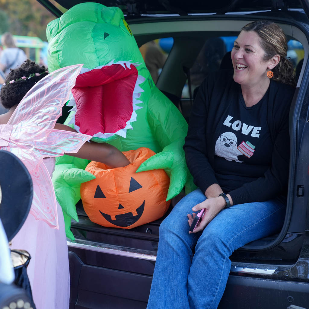 A child in pink fairy wings reaches into an inflatable jack-o’-lantern candy bowl held by a green monster while a seated adult smiles nearby.