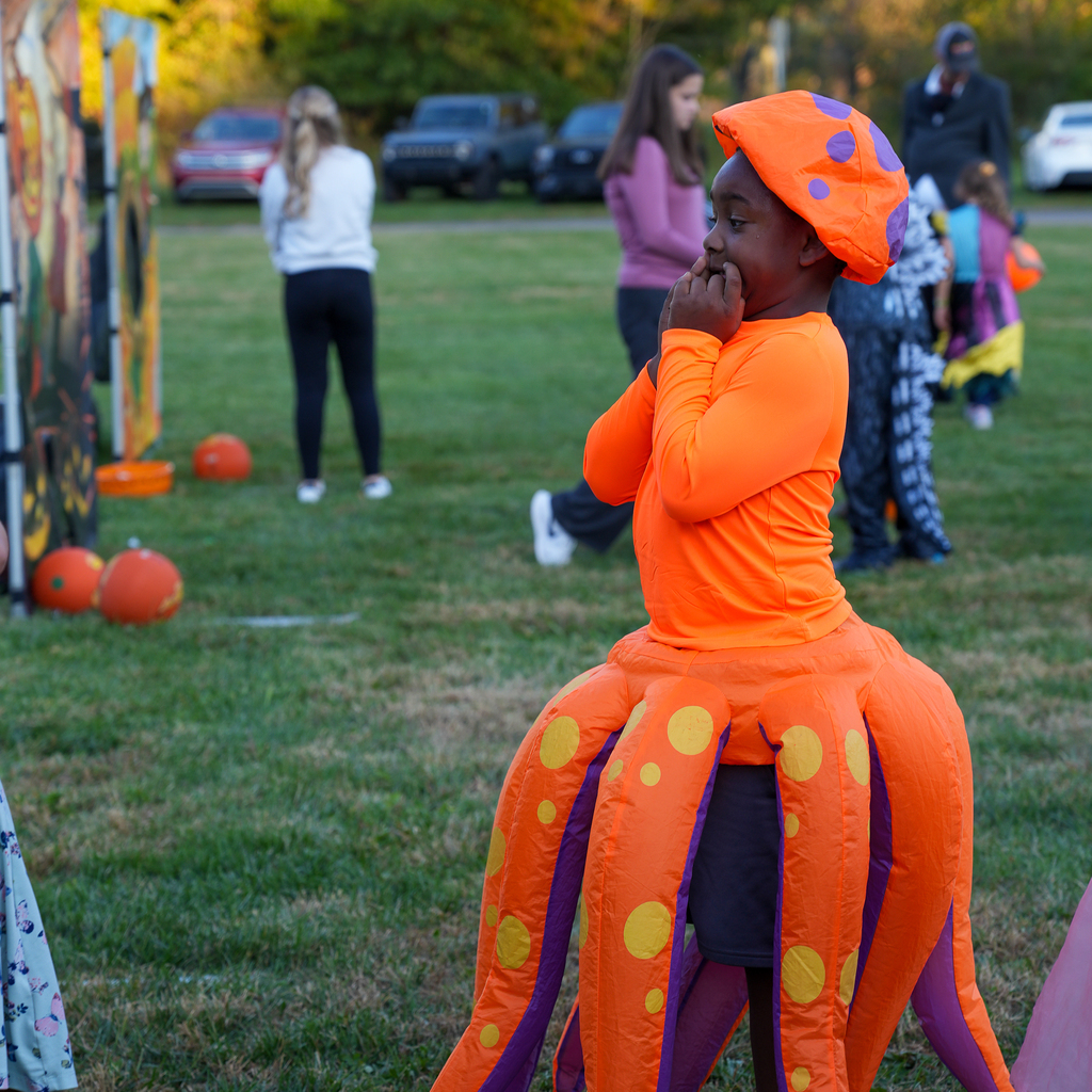 A child in a bright orange and purple octopus costume holds their hands near their mouth while looking toward a festival activity.