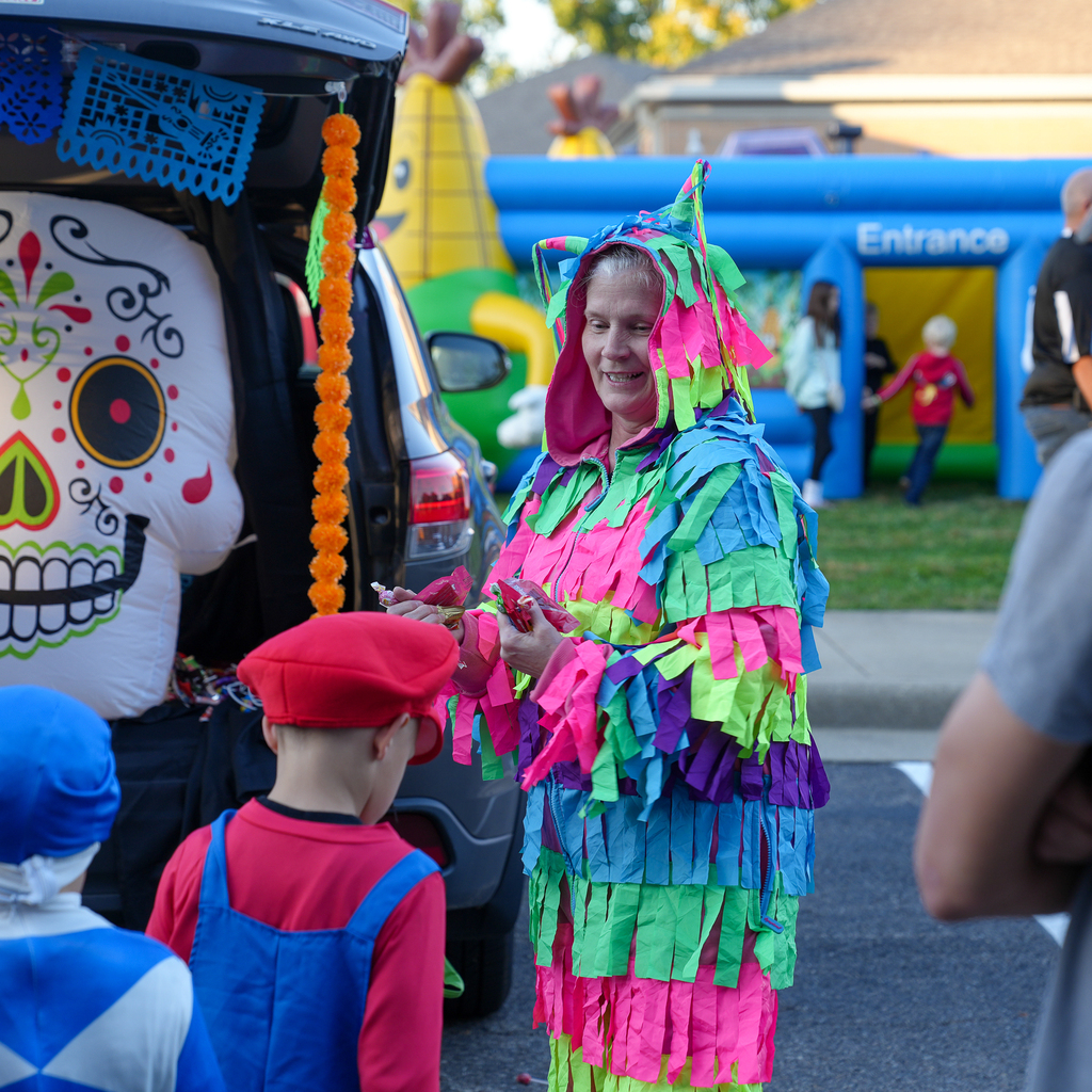 An adult in a colorful piñata costume holds candy for children dressed as Mario and a blue Power Ranger beside a decorated vehicle.