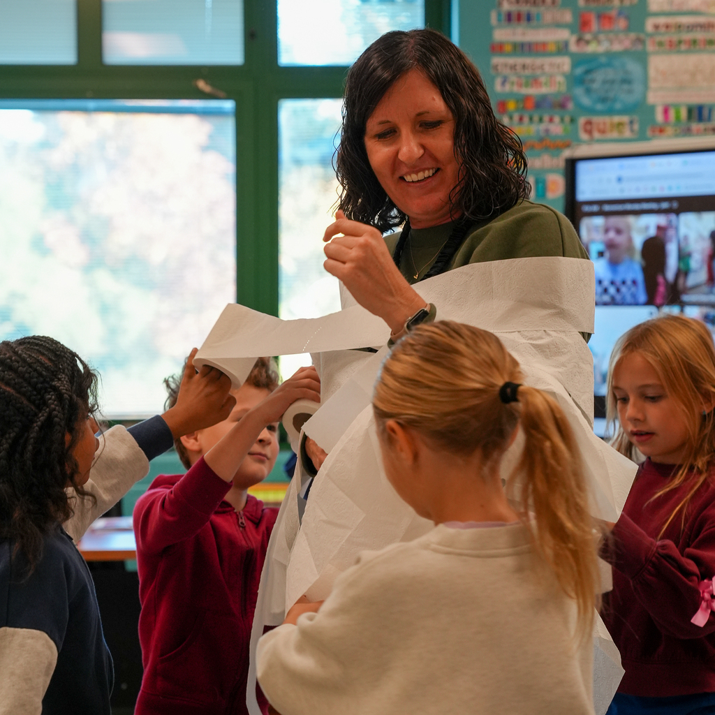 Students work together to wrap a smiling staff member’s arms and torso in toilet paper.