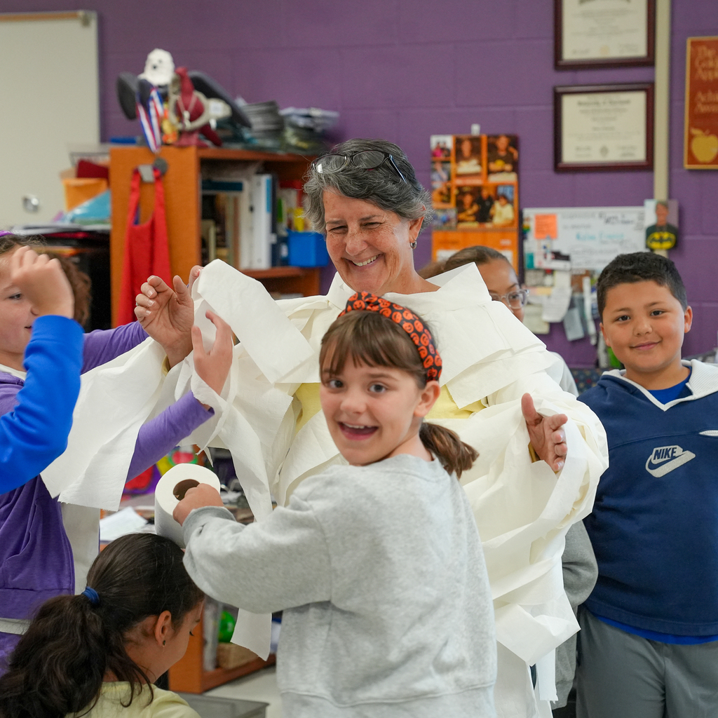 A smiling staff member holds their hands up as students add layers of toilet paper to their mummy costume.