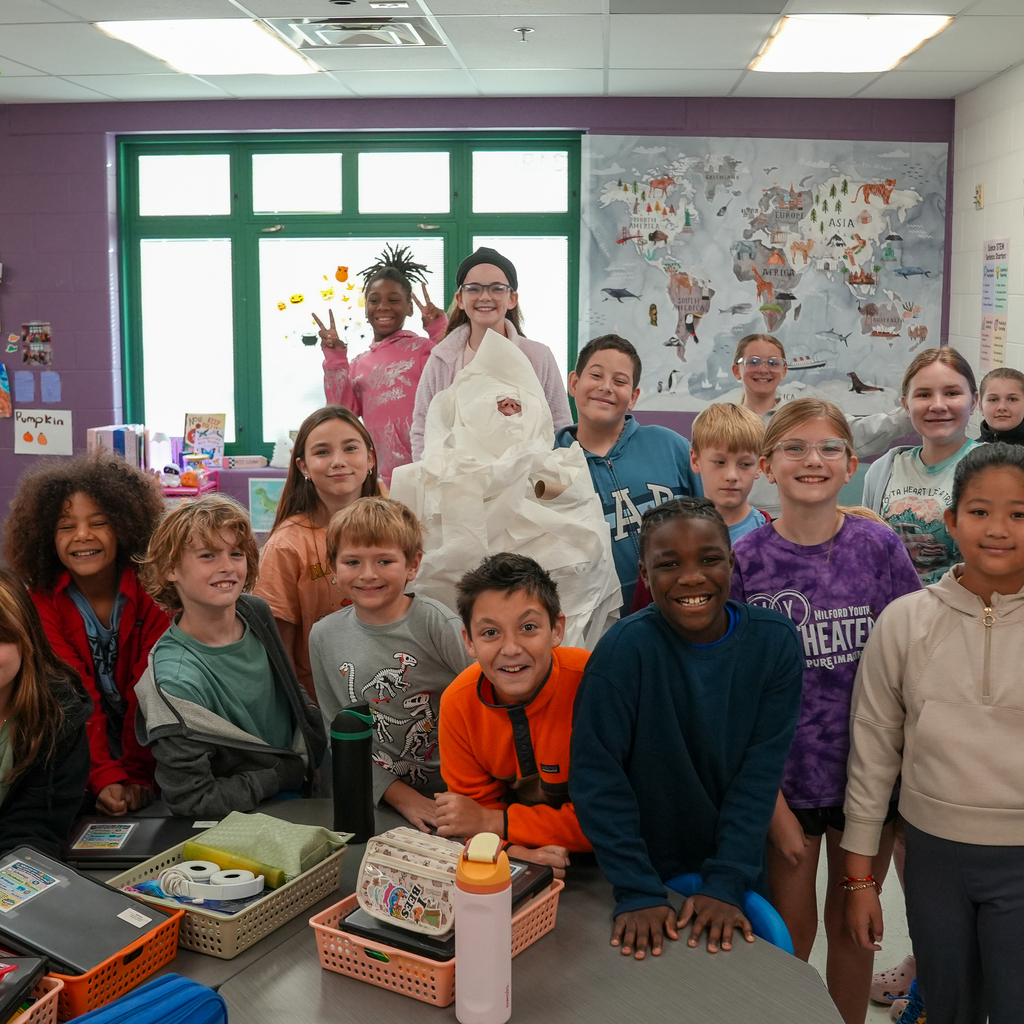 Students smile and pose around a person wrapped in white toilet paper like a mummy in a Mulberry classroom.