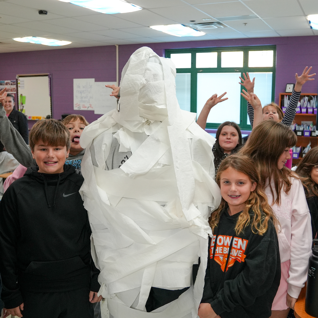 Students pose beside a person covered from head to legs in layers of white toilet paper, with classmates raising their hands behind them.