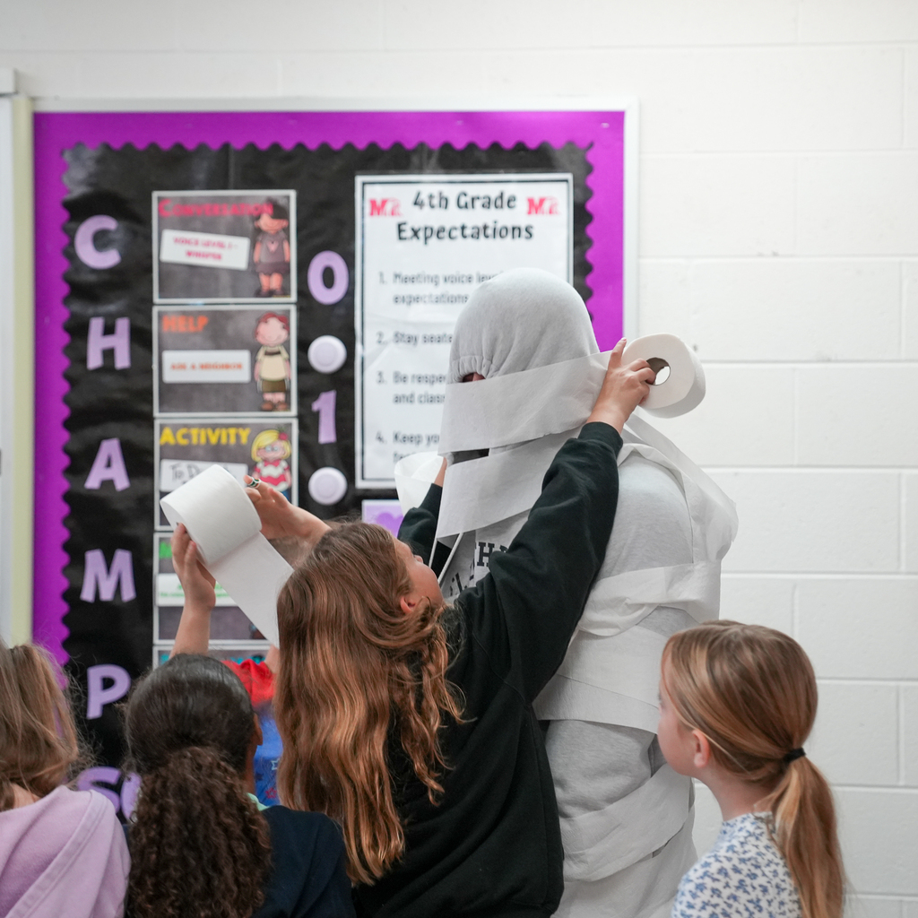 Students reach up with toilet paper rolls to wrap a hooded staff member’s head and shoulders during a classroom mummy activity.