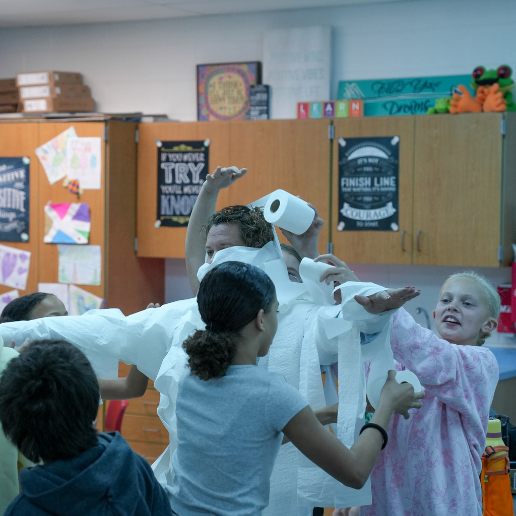 Students wrap a staff member in toilet paper as the staff member extends both arms outward.