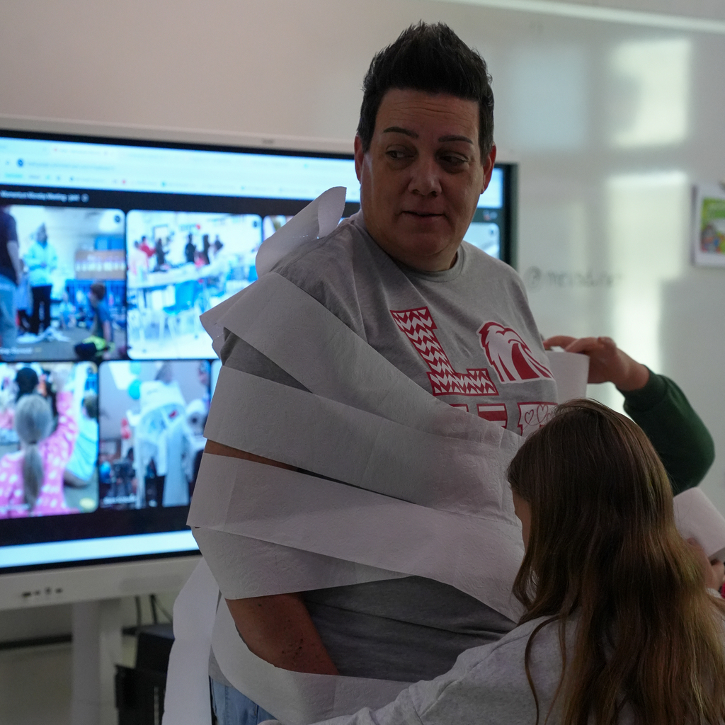 A staff member looks toward students as they wind strips of toilet paper around the staff member’s torso and arms.