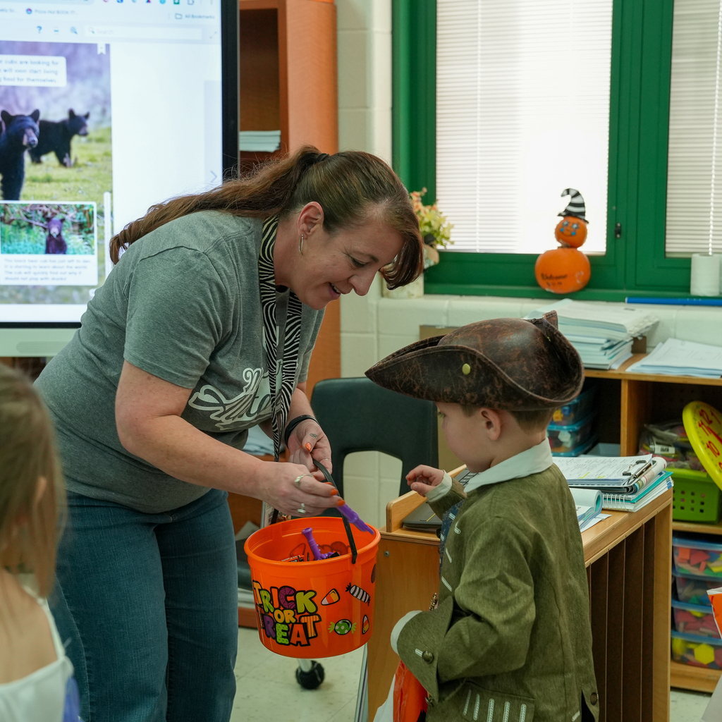 A staff member offers a treat from an orange Halloween bucket to a preschooler wearing a pirate hat and green coat.