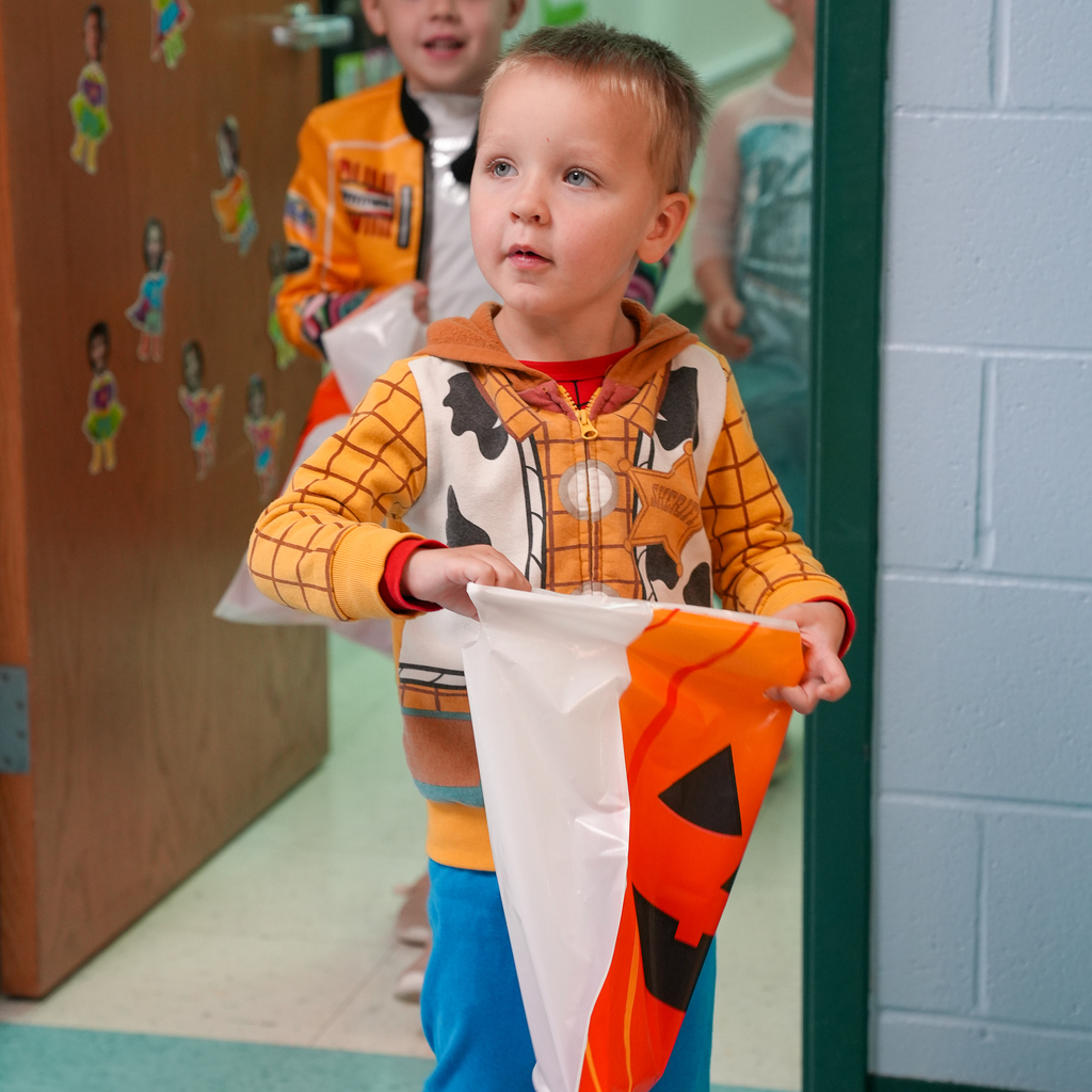 A preschooler wearing a Woody-themed cowboy costume holds a jack-o’-lantern treat bag in a classroom doorway.