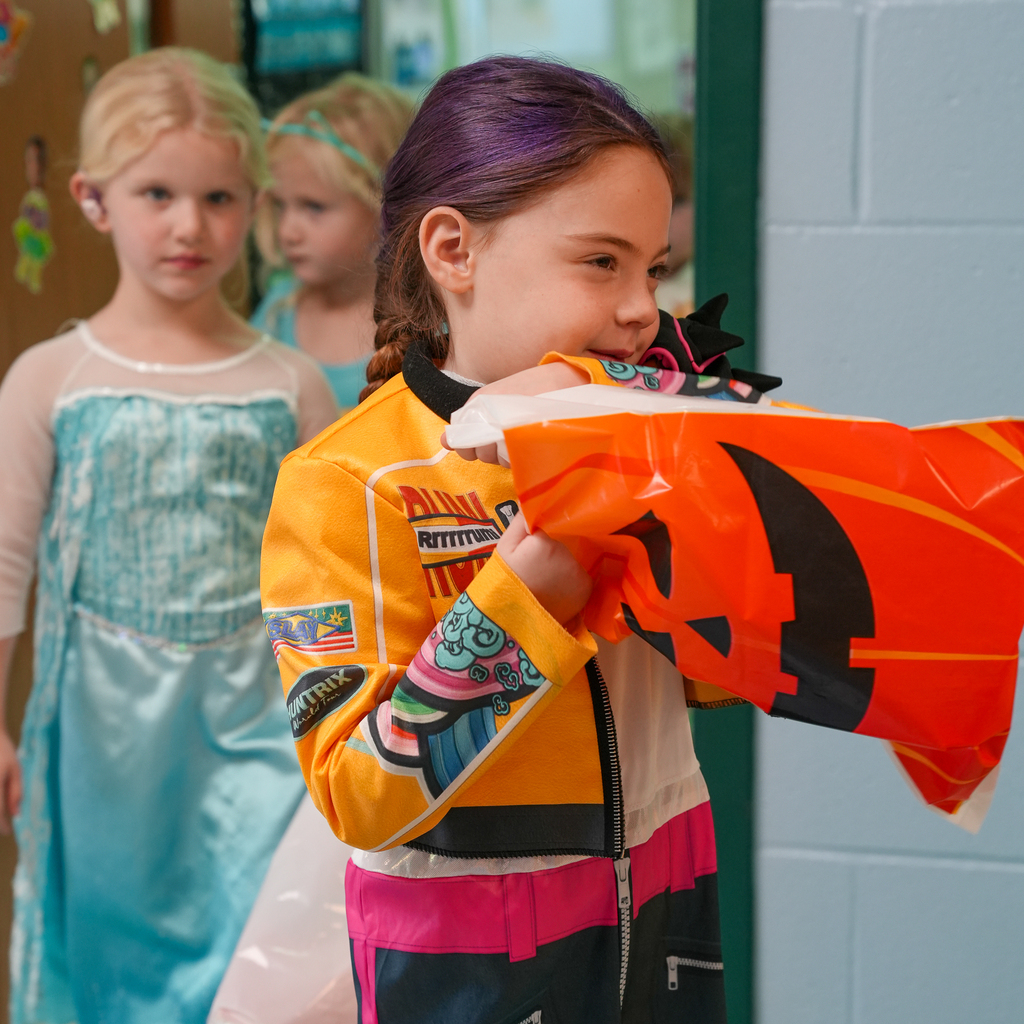 A preschooler with purple-colored hair and a bright racing costume holds up a jack-o’-lantern treat bag while children in princess dresses wait behind.