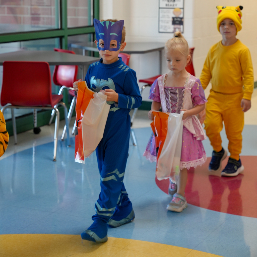 Preschoolers dressed as Catboy, a princess and Pikachu walk through school, carrying bags for trick-or-treating.