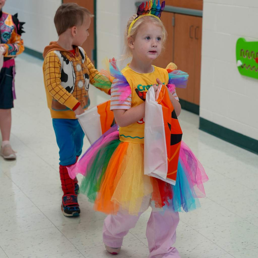 A preschooler wearing a rainbow tutu and crayon headband carries a jack-o’-lantern treat bag down the hallway, followed by costumed classmates.