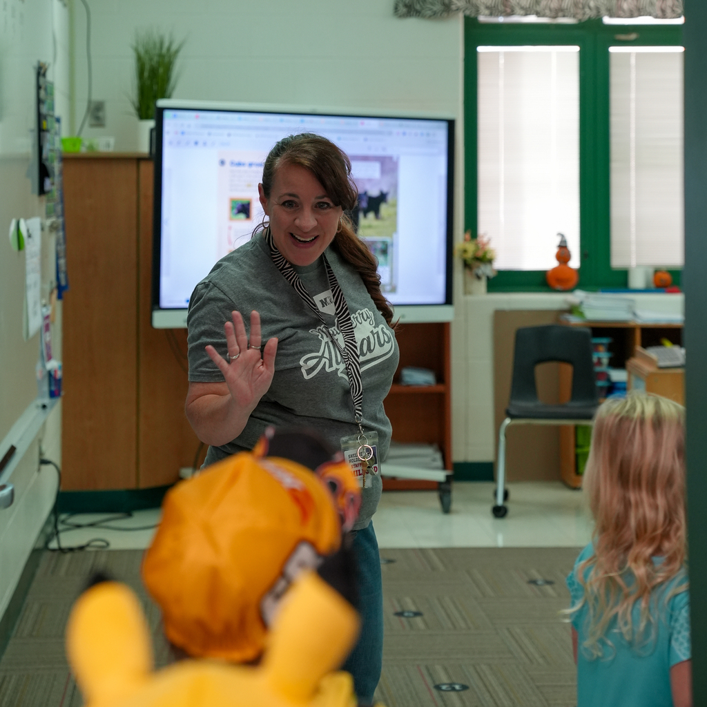 A smiling staff member waves to costumed preschoolers visiting a classroom during trick-or-treating.