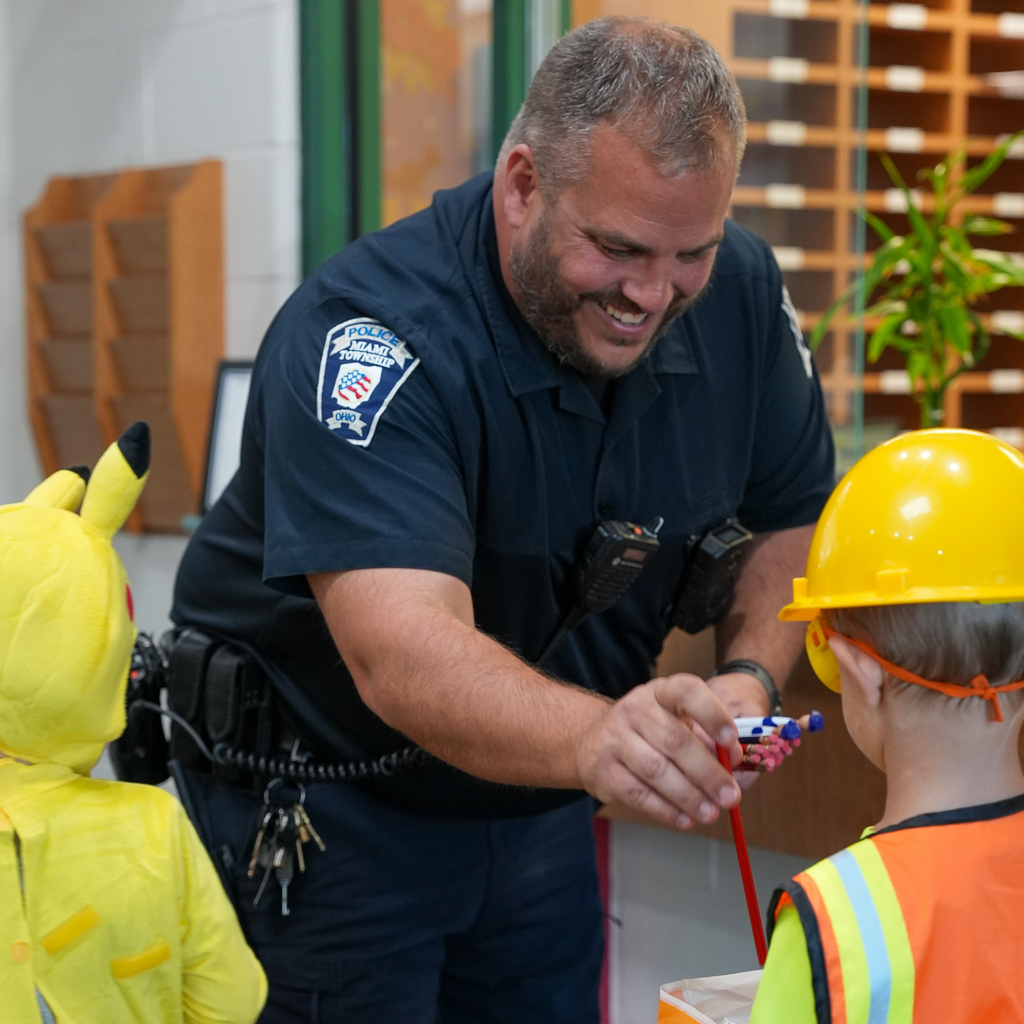 A smiling Miami Township police officer offers a pencil to a preschooler wearing a yellow construction helmet and orange safety vest.