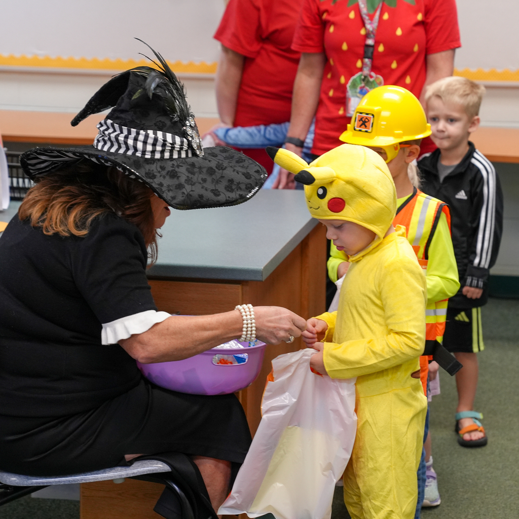 A staff member wearing a witch hat hands a treat to a preschooler dressed as Pikachu while other children wait in line.