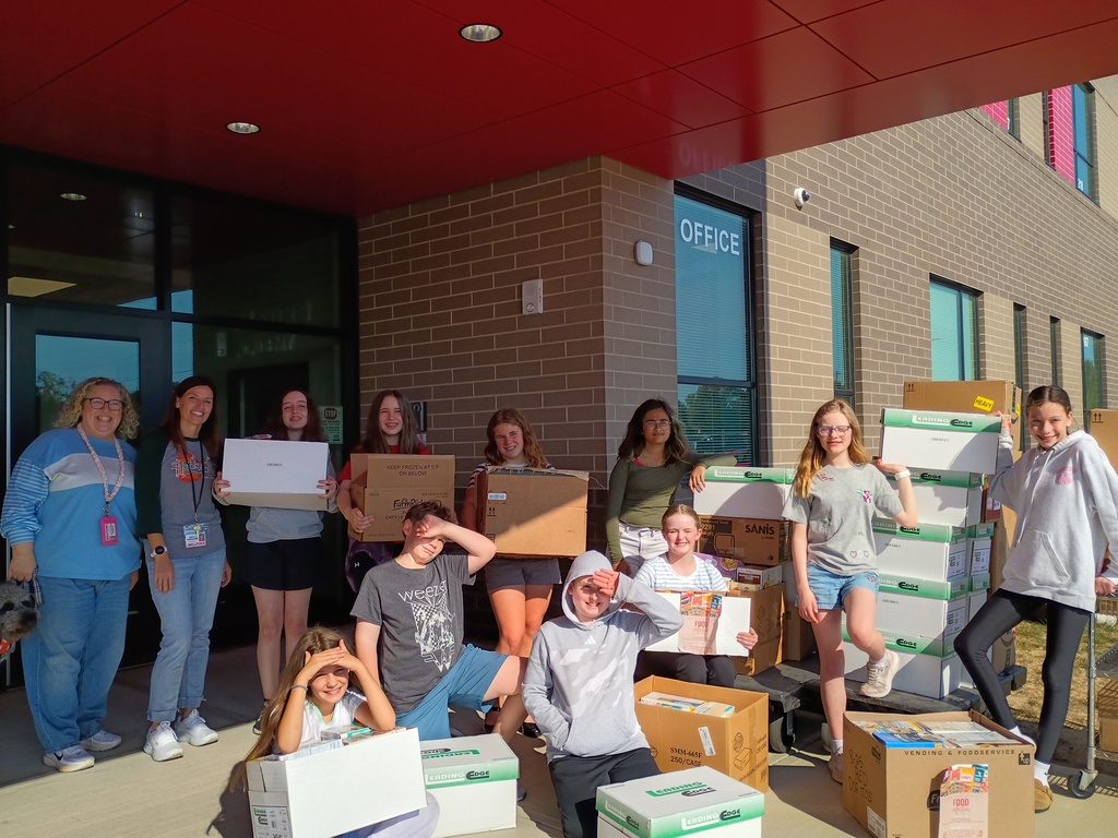 Students and two adults pose outside a school entrance with stacks of cardboard boxes, several filled with packaged food and labeled for a food drive. Some hold boxes while others kneel in front, shading their eyes from the sun.