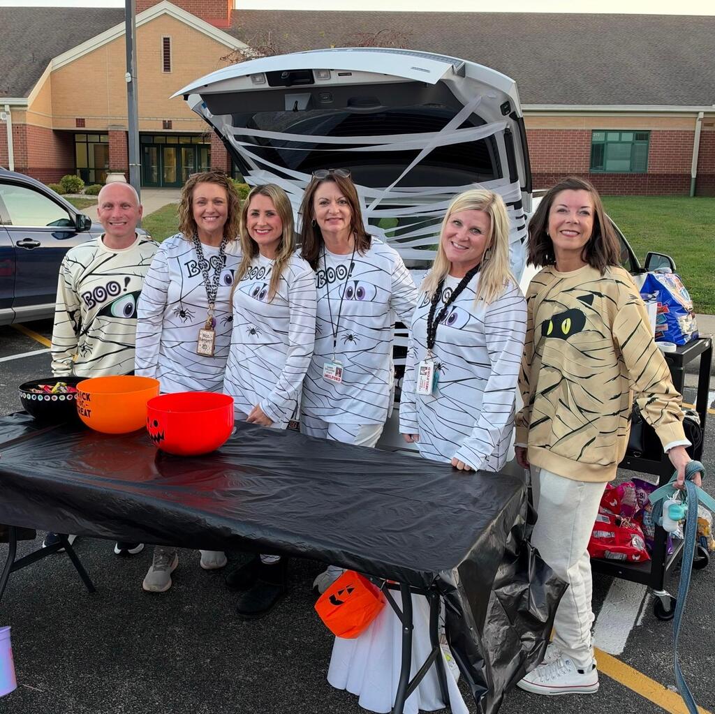 Six staff members wearing mummy-themed shirts smile behind a table with Halloween candy bowls. Behind them, an open vehicle trunk is decorated with white streamers resembling mummy wrappings outside a school building.