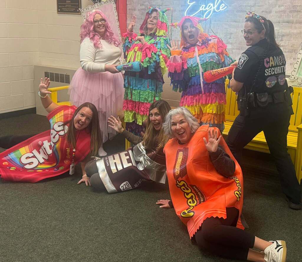 Seven staff members pose playfully in candy-themed Halloween costumes, including Skittles, a Hershey’s bar, Reese’s, two colorful piñatas, a pink-haired character and a black outfit labeled “Candy Security.” Several wave at the camera while three sit on the floor in front.