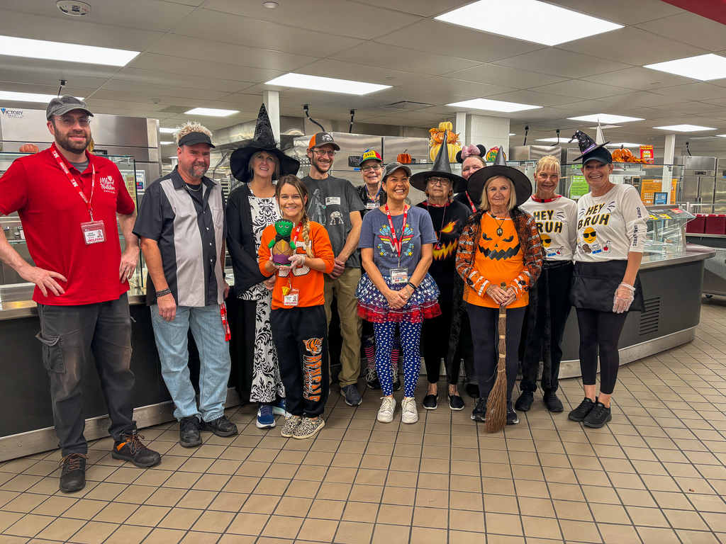School nutrition staff pose in the cafeteria wearing Halloween costumes and festive clothing, including witch hats, pumpkin shirts and a Wonder Woman outfit. One holds a Frankenstein figure, and another holds a broom.