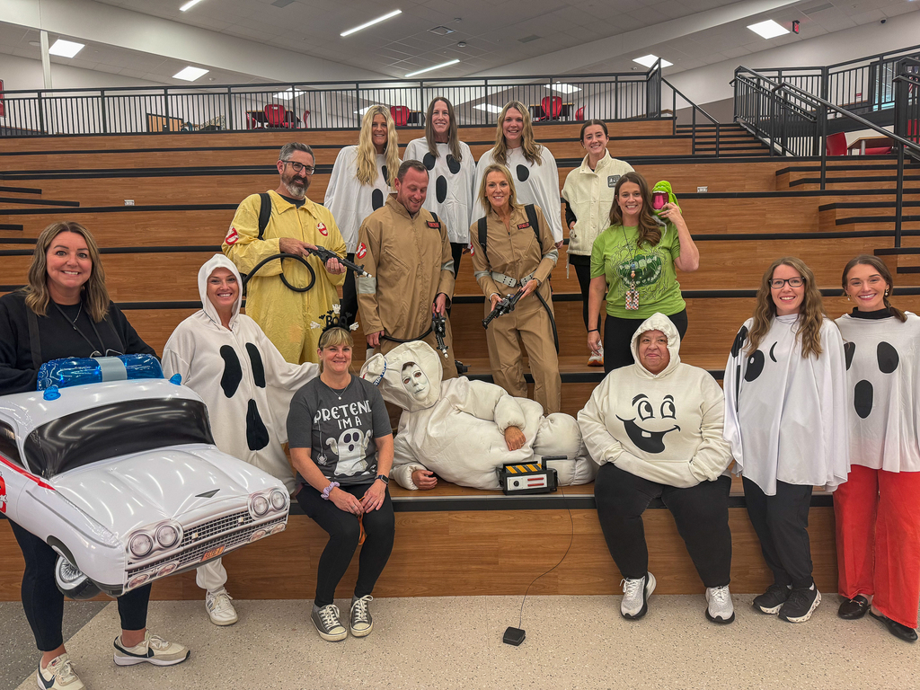 Staff members pose on tiered wooden seating in Ghostbusters-themed costumes, including jumpsuits with prop equipment, white ghosts, Slimer and the Stay Puft Marshmallow Man. An inflatable Ghostbusters car is at the front.