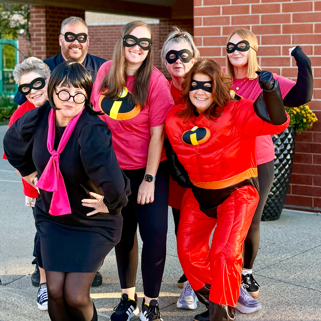 Seven staff members pose outside a brick school building dressed as characters from “The Incredibles,” including Edna Mode and masked superheroes. Several flex their arms or stand with hands on their hips.