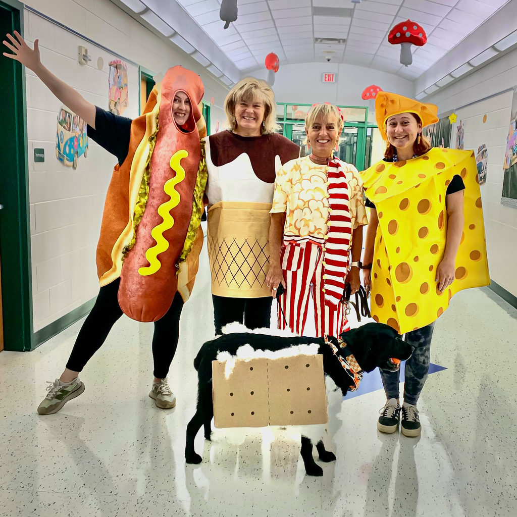 Four staff members smile in a school hallway dressed as a hot dog, ice cream cone, popcorn and cheese. A black dog wearing a s’more costume stands in front of them.
