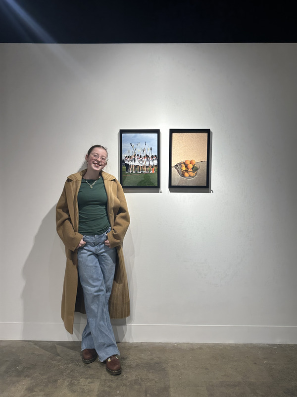 A smiling young woman in a tan coat and jeans stands in an art gallery beside two framed photographs—one of a lacrosse team celebrating and one of oranges in a bowl.
